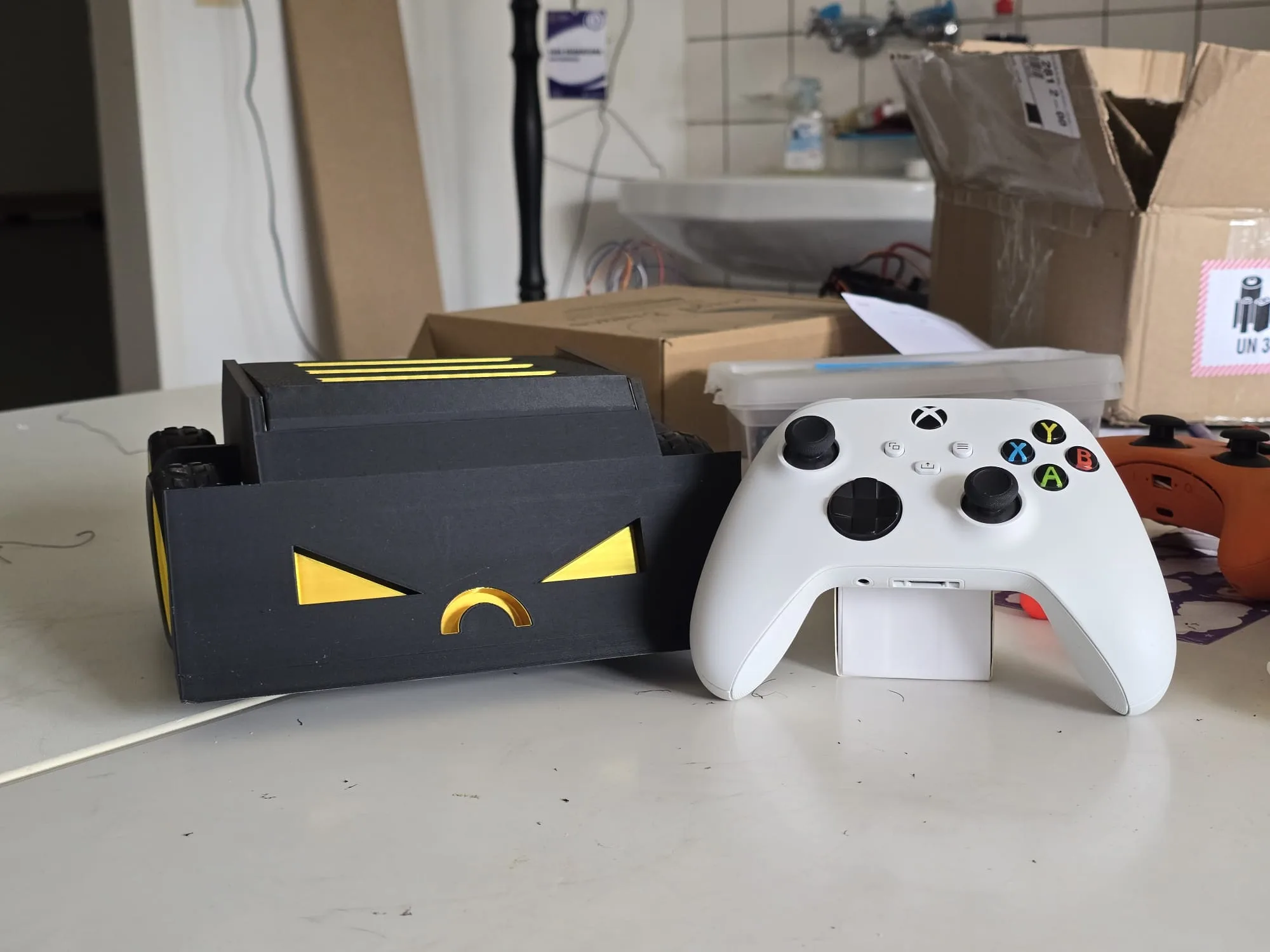 Sumo robot with a black chassis and yellow eye markings, beside the Xbox controller that drives it